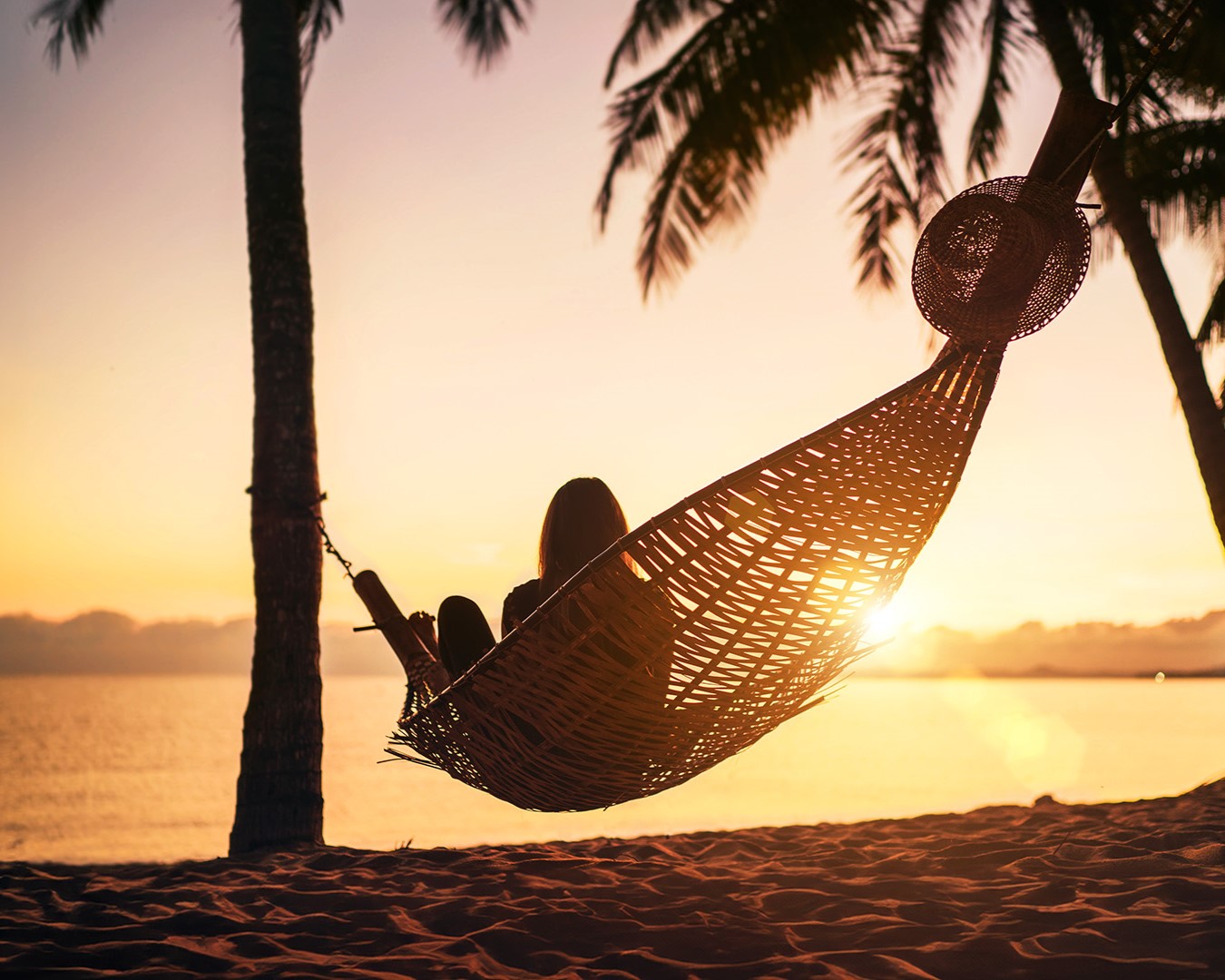 Frau in Hängematte am Strand beim Sonnenuntergang, entspannter Urlaubs-Moment am Meer