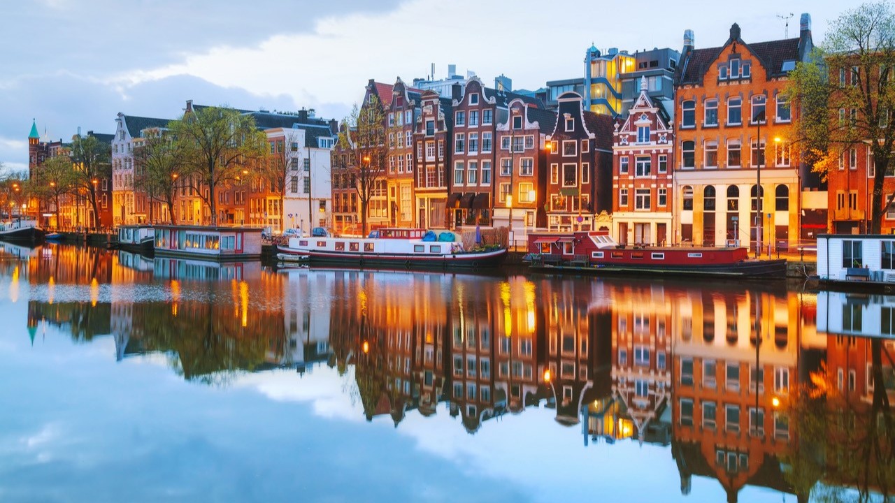 Blick auf beleuchtete Häuser am Wasser in Amsterdam bei Dämmerung, historische Gebäude entlang der Grachten | © Andrey Krav, iStock