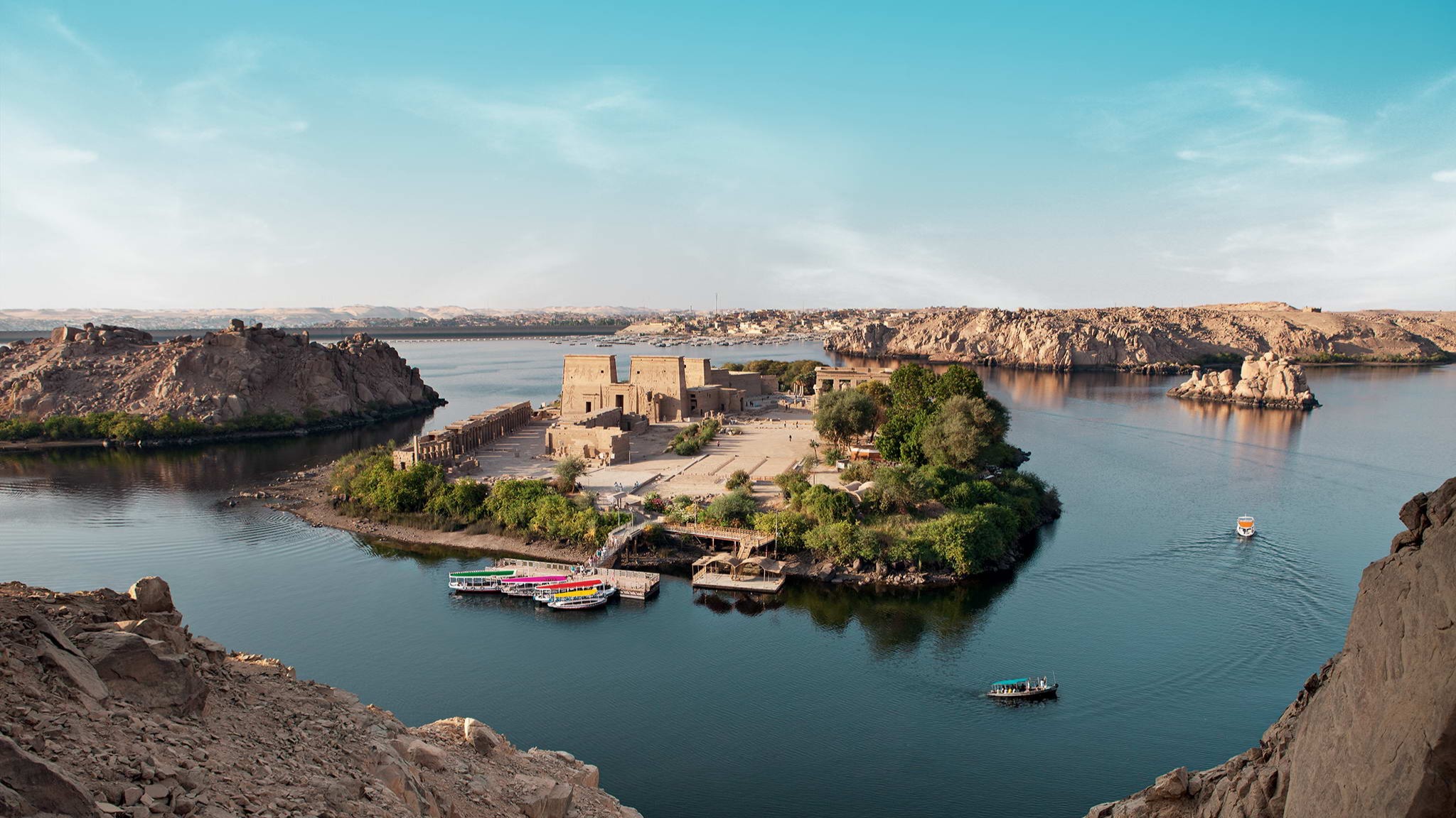 Luftaufnahme einer kleinen Insel im Nil mit alten ägyptischen Tempelruinen, umgeben von ruhigem Wasser. Mehrere bunte Boote liegen am Ufer, im Hintergrund sieht man felsige Uferlandschaften und die weitläufige Flusslandschaft. | © DERTOUR