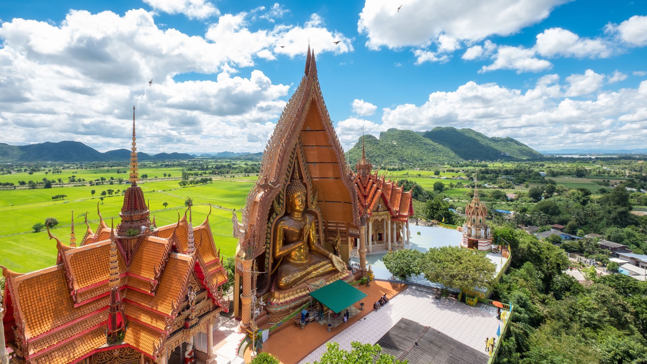 Luftaufnahme des Wat Tham Sua Tempels in Kanchanaburi, Thailand, mit einer goldenen Kirche, einer großen Buddha-Statue und umliegenden grünen Reisfeldern | © Getty Images